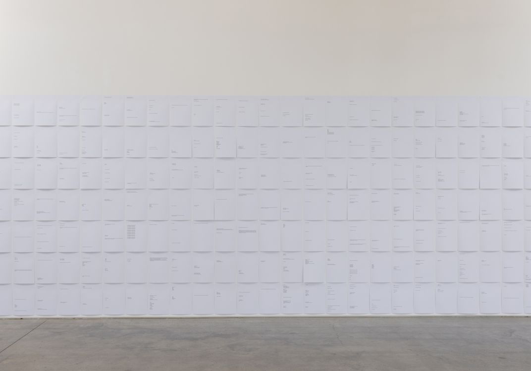 Wall covered with white paper sheets arranged in a grid, each displaying minimal printed text; bare concrete floor in foreground.