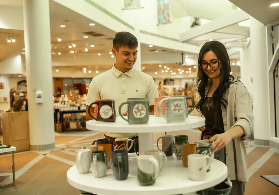 Visitors examine handmade pottery displayed inside a spacious craft gallery. Shelves and displays throughout the marketplace showcase a variety of locally made arts and crafts.
