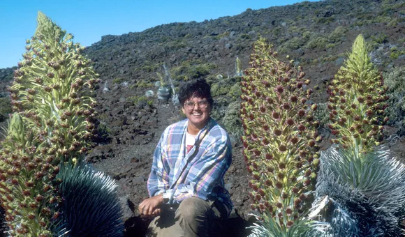 A woman in a blue checkered shirt kneels on the ground with tall, green plans surrounding her on both sides.