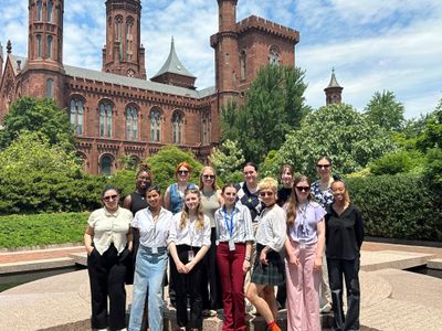 A group of thirteen people stand together in front of green shrubs and the Smithsonian Castle.