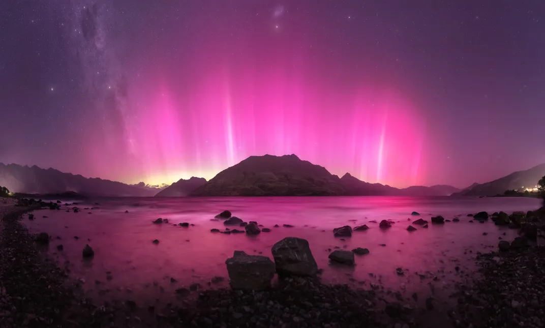 A pink aurora shines over a mountain and water in the night sky, with streaks emanating up from the horizon like fire