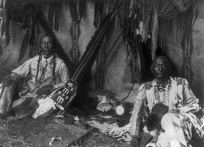 Yellow Kidney (left) and his father, Little Plume, inside a lodge, pipe between them.