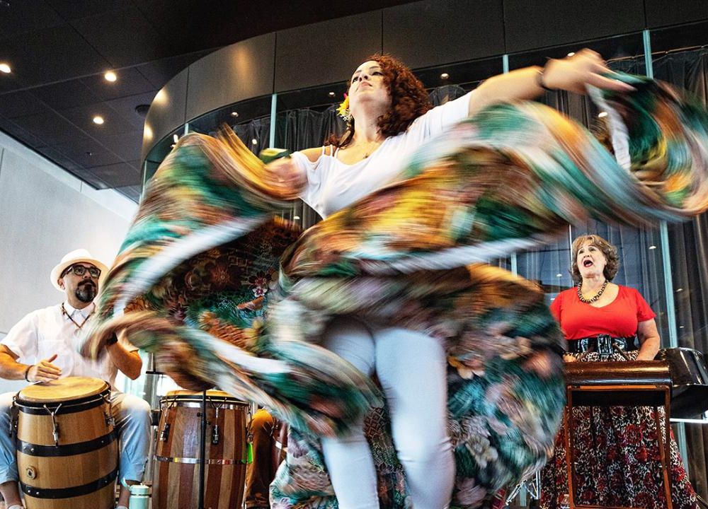 A person dances, turquoise and brown skirt flying and twisting around them, with a drummer and singer performing on the sides.
