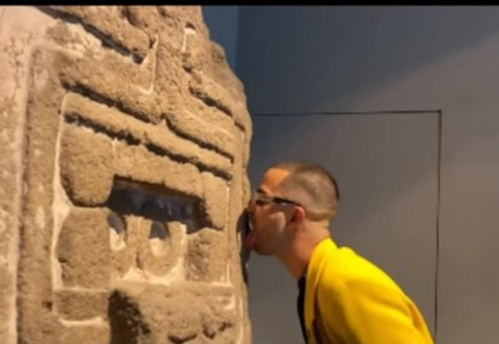In a screenshot, a man in a yellow jacket licks the carved stone facade of an ancient work of Indigenous art