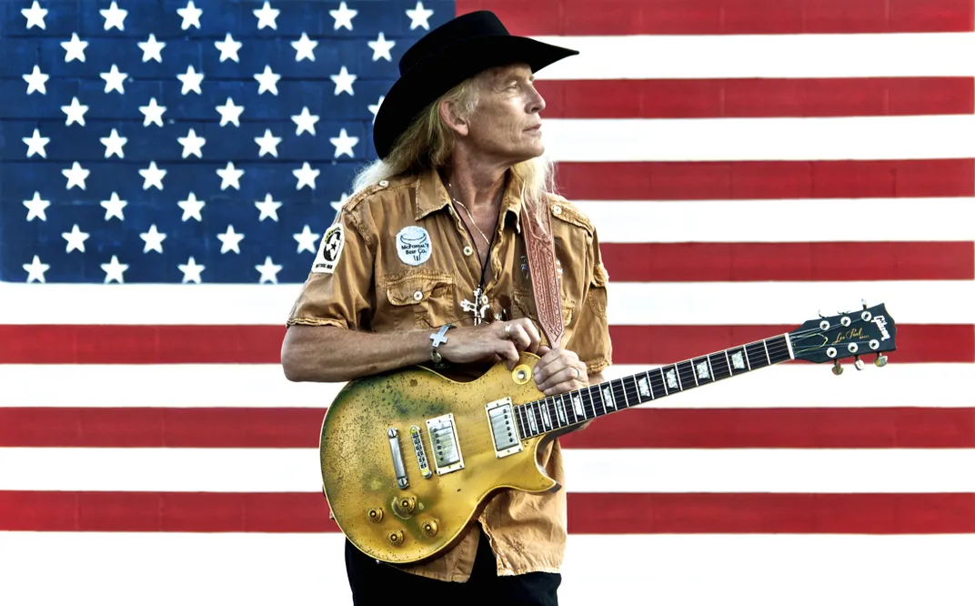 A older man stands with a guitar in front of the American flag