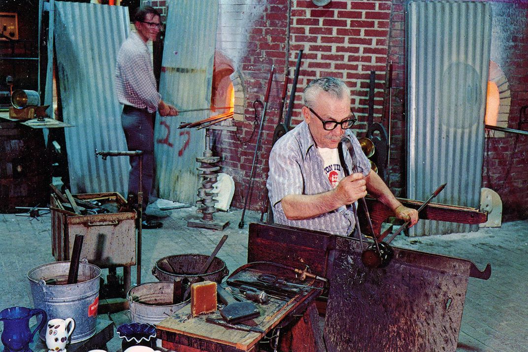 In a brick-walled factory, a man sits and uses two long metal tools to manipulate a dark, round object. Another man in the backgrounds holds a long metal rod in a glowing furnace.