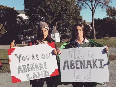 Two protesters hold signs saying "You're on Abenaki land" and "This is Abenaki land."
