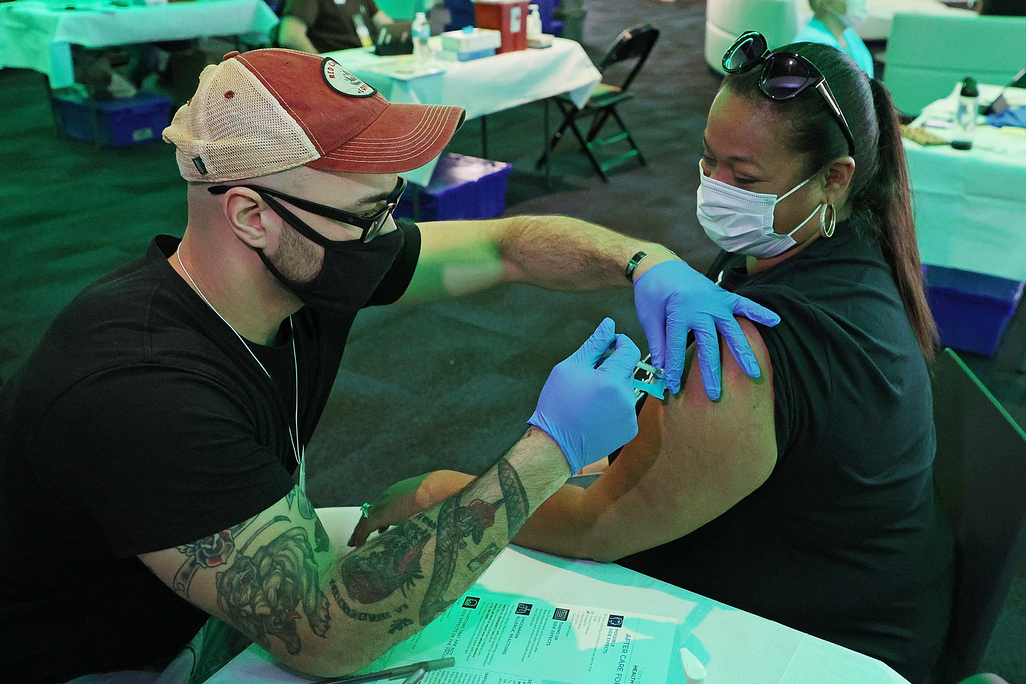 A woman in a black shirt and sunglasses gets a vaccine from a man in a black shirt and red baseball cap, both sitting at a white table and wearing masks