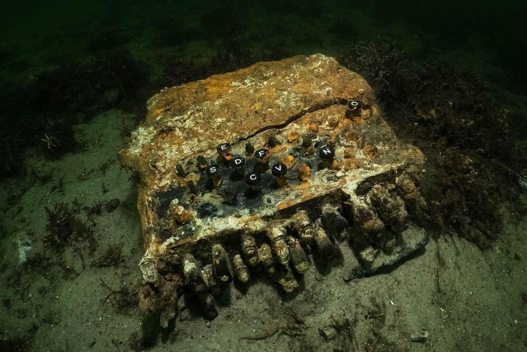 A close-up shot of the rusted machine at the bottom of the ocean, a bit overgrown with orange-y algae but with its keys, like a typewriter's, still discernable