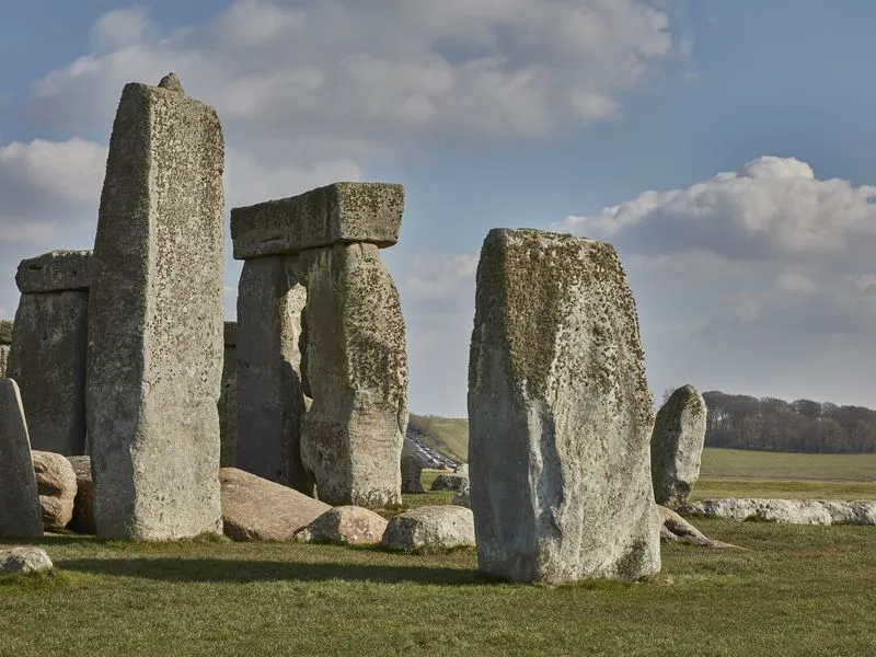A view of the iconic rocks of Stonehenge on green grass, with a sloping hill visible between two stones in the distance and a line of traffic
