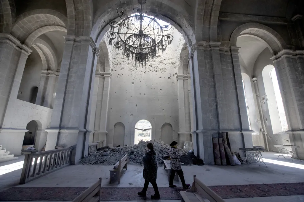 Two people walk, cast in shadows, underneath the tall white arches of a cathedral; behind them, a pile of rubble blocks a doorway and a broken chandelier hangs from the ceiling