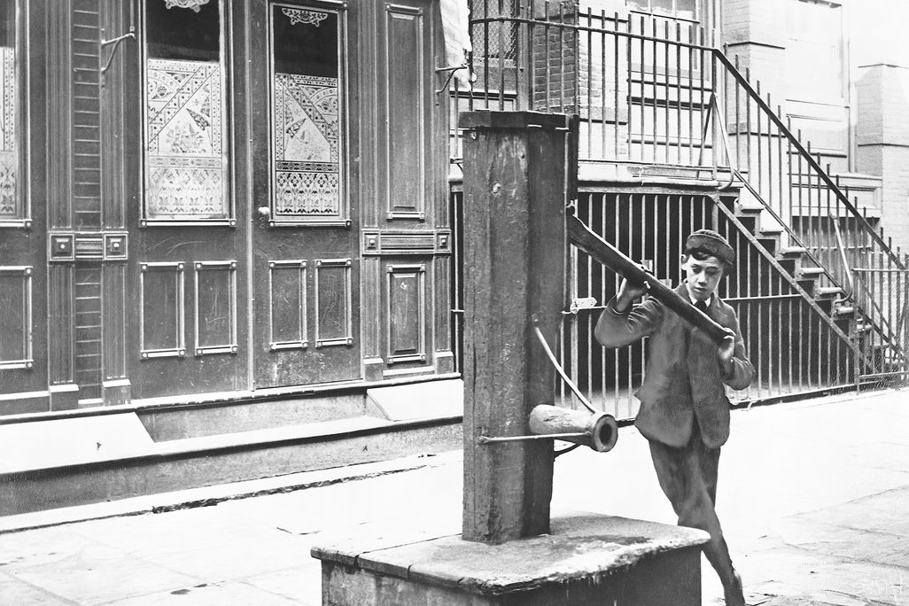 An old photo of a boy holding the handle of water pump on a street in New York City