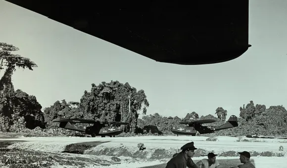 Three men sit under the shade of a massive airplane wing; one of the men is wearing an officer's hat. Two huge aircraft are parked in the background, both are painted black. Behind them is a lush island landscape. The photo has been given a green tint.