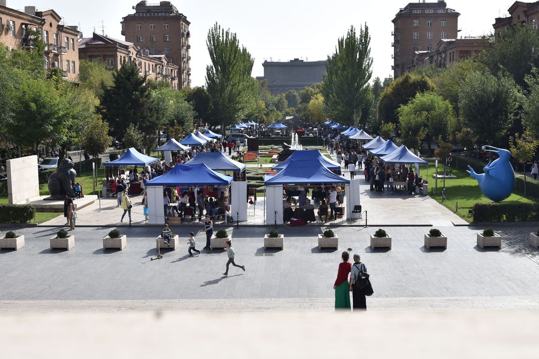 A large, rectangular sculpture park is filled with blue tents for vendors. In front of the tents is a patio area where children play.