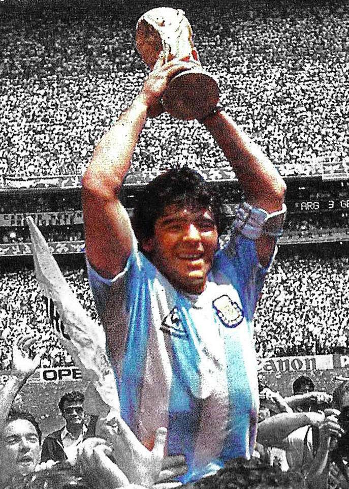 Maradona, in blue and white jersey, is hoisted up in the air by other people, and he holds up a trophy. Enormous crowd in a stadium behind him. He is in color, and everything else in black and white.