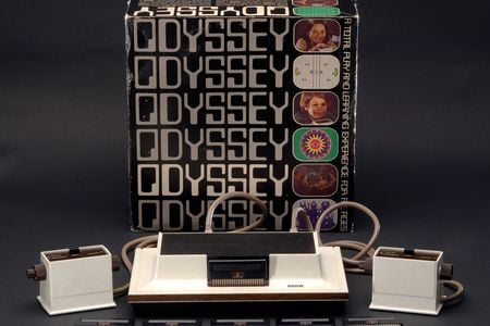 The Magnavox Odyssey system arrayed in front of its cover box. The box is decorated with the system's name and small images of various games and children using the Odyssey's controller