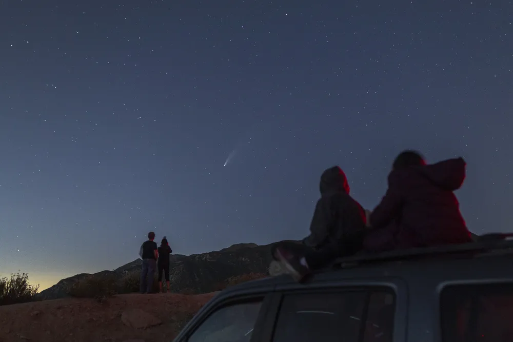 Outlines of figures, including two young people seated on top of a car, framed against twilight sky as they watch a comet streak across the sky
