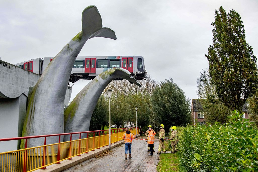 A red and silver metro train rests precariously, hanging off of the train tracks, with the first car resting on one of two enormous sculptures of whale tails. Below, six construction workers asses the damage.