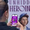 View from the back as a woman pulls up a webpage on her phone. In the background a poster reading “Unhidden Heroines” is visible.