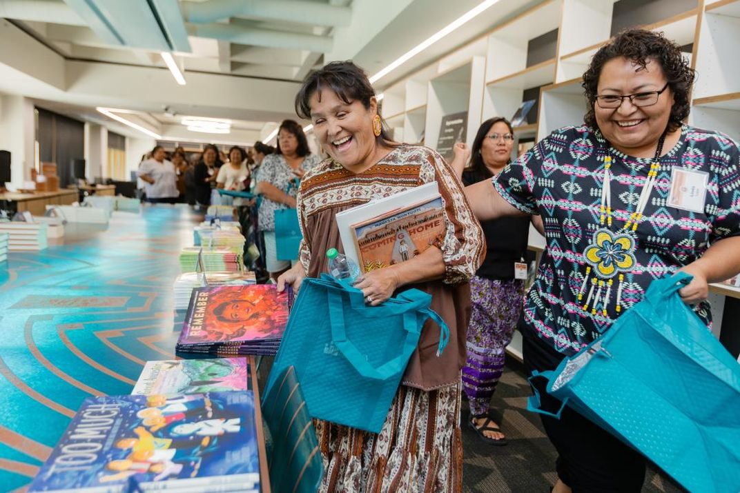 Two women laugh as they pick up books. There is a line behind them.