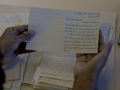 Two hands holding a card with handwritten information. A stack of other handwritten papers is visible on the table below.