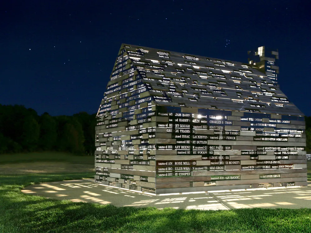 A cabin structure is illuminated from within, with light pouring out in between metal slats engraved with lines of poetry