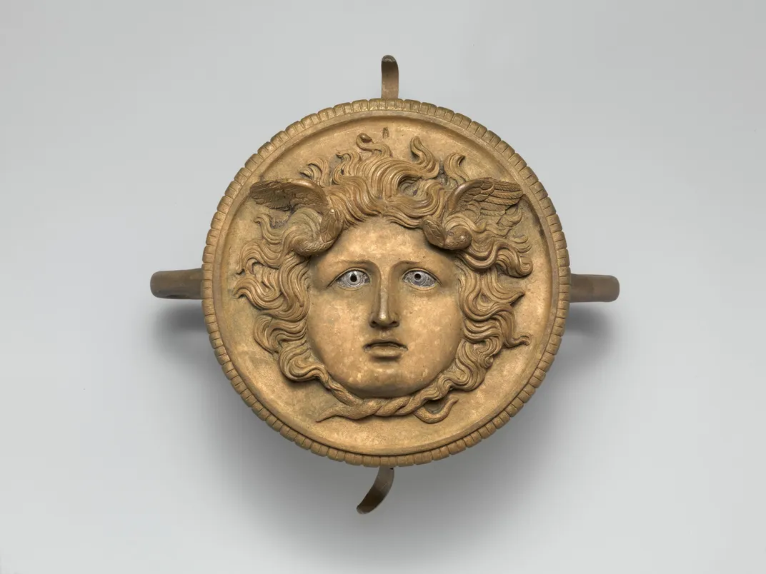 A yellowish circle with a carved face of Medusa, a beautiful female face with flowing hair encircling her head and small wings extending like antlers