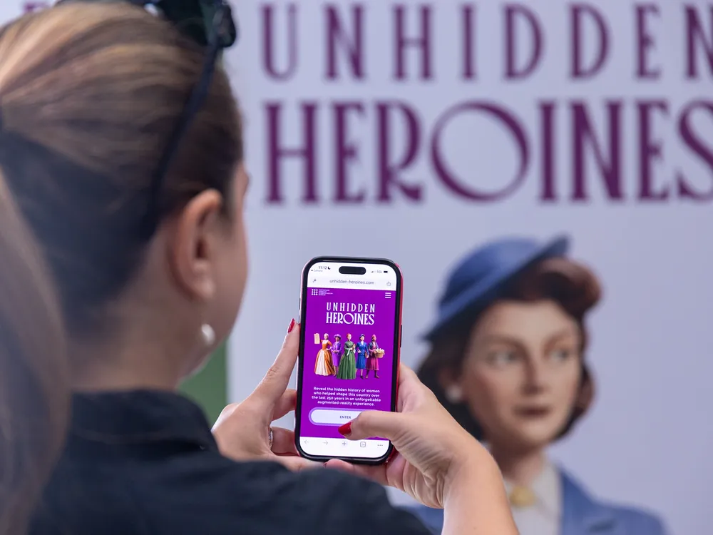 View from the back as a woman pulls up a webpage on her phone. In the background a poster reading “Unhidden Heroines” is visible.