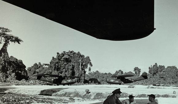 Three men sit under the shade of a massive airplane wing; one of the men is wearing an officer's hat. Two huge aircraft are parked in the background, both are painted black. Behind them is a lush island landscape. The photo has been given a green tint.