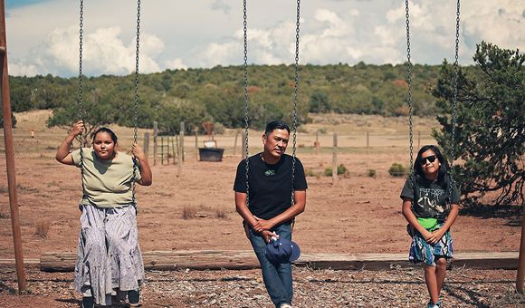 Two children and an adult sit on three swings in a dry landscape with trees in the background.
