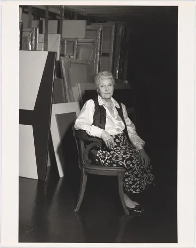 A black and white photograph of a woman sitting in a chair in front of canvases.