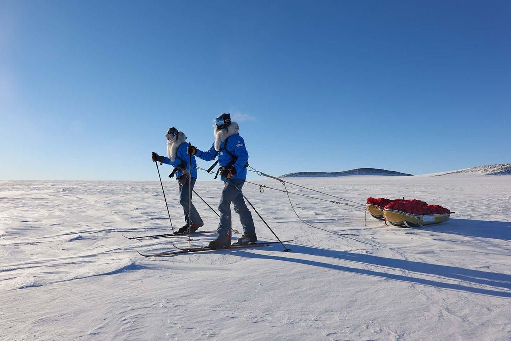 Two men in winter clothes and skis pull sleds across and frozen landscape