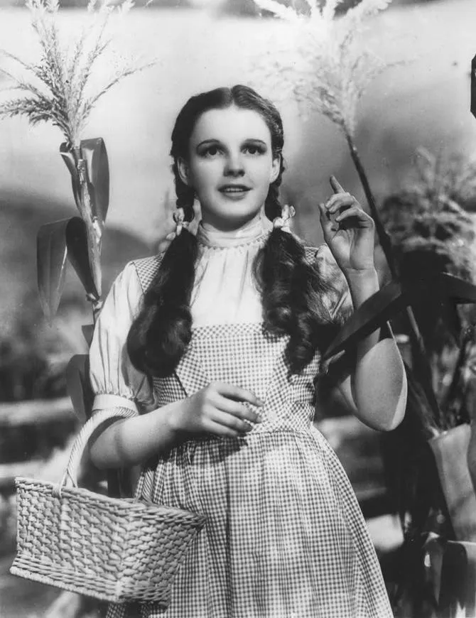 A black and white image of a young white girl, Garland, with dark hair in two braids, carrying a basket and wearing a checked pinafore over a white shirt