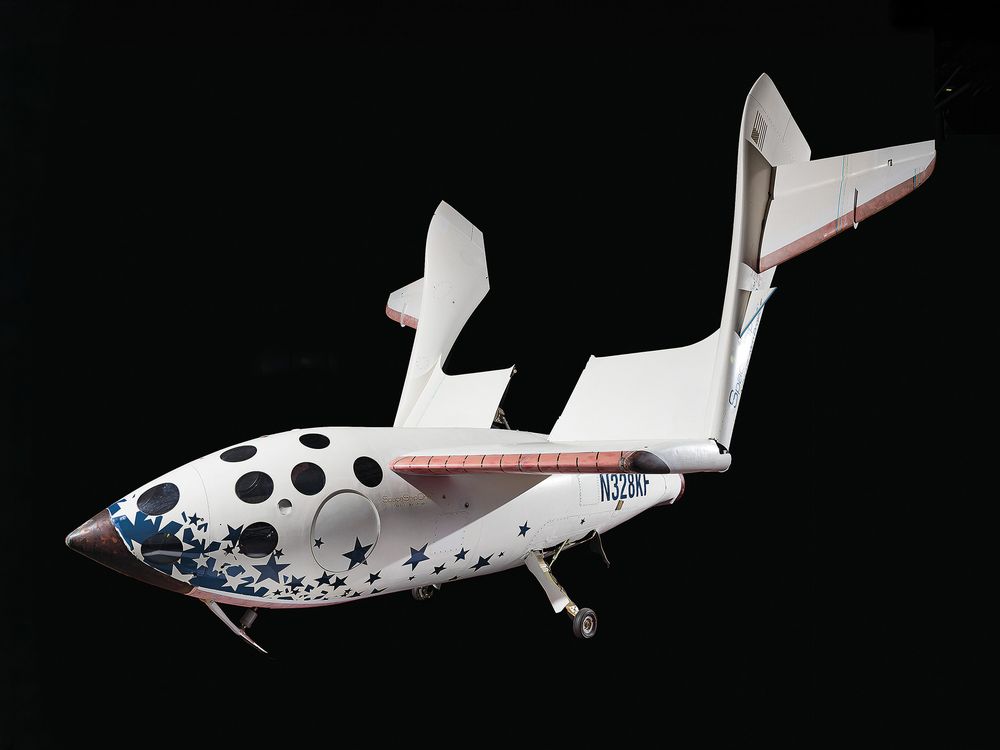 A white spacecraft against a black background. The fuselage is spangled with dark blue stars and black circles. A large wing is swiveled up away from the fuselage. The tail of the spacecraft is marked by this identifying number: N328KF.