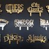 A collection of twelve custom-made, metallic signs is displayed against a dark background. The signs are arranged in three rows of four. The signs are a mix of gold and silver colors with various fonts and designs.
