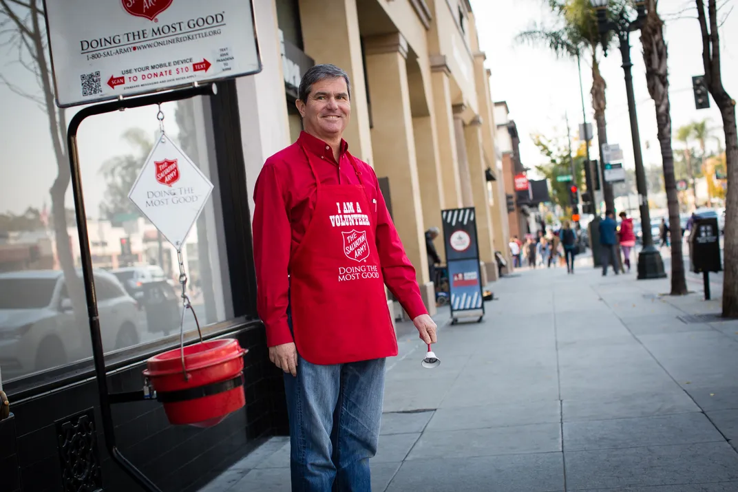 Man wearing a red apron holding a bell and standing in front of a red kettle