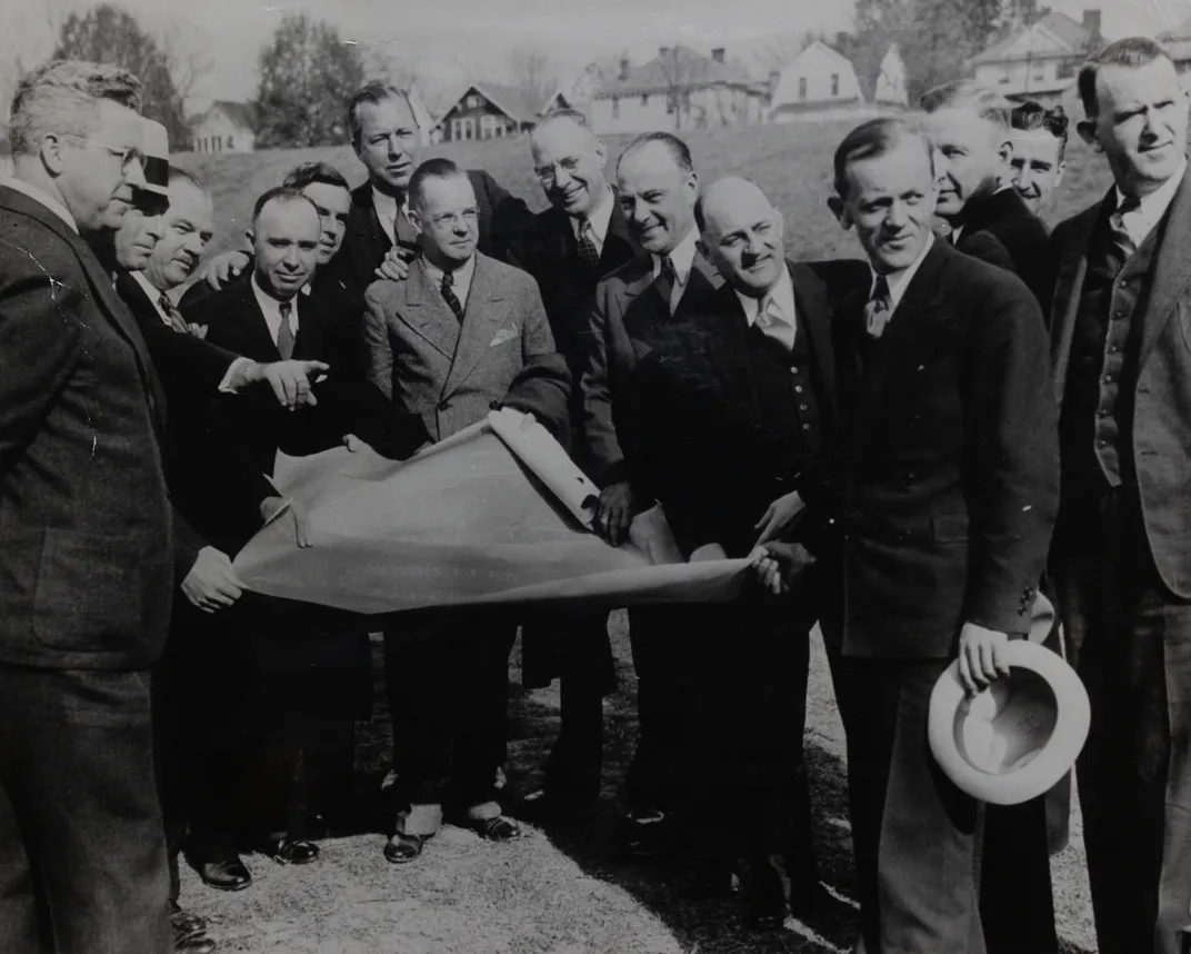 A black and white photograph of a group of white men in suits holding a large blueprint