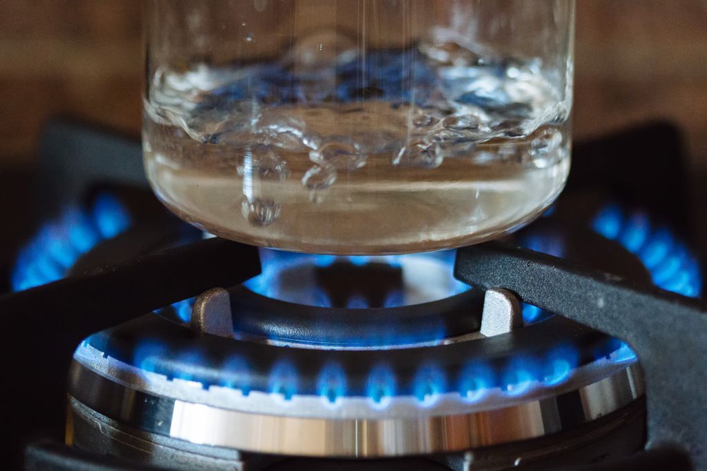 A close-up of a blue-flame burner on a stove, with water boiling in a clear glass pot.
