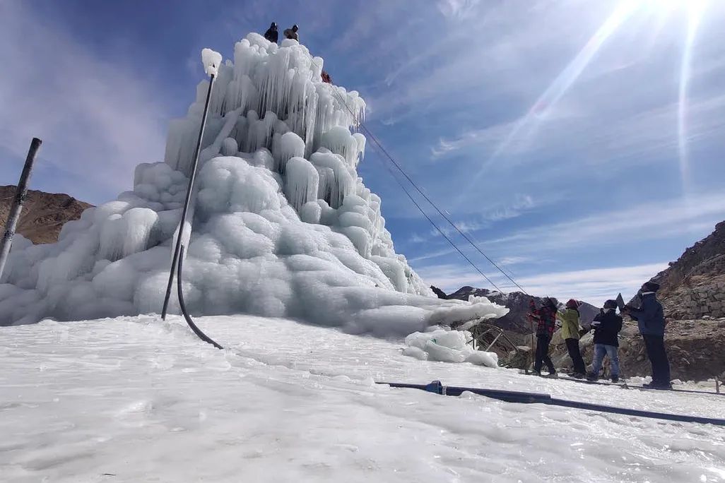 On a bright day in the Ladakh Valley, a team of people work on an ice stupa. It is a tall, cone-shaped pile of ice that sits upon snowy ground.