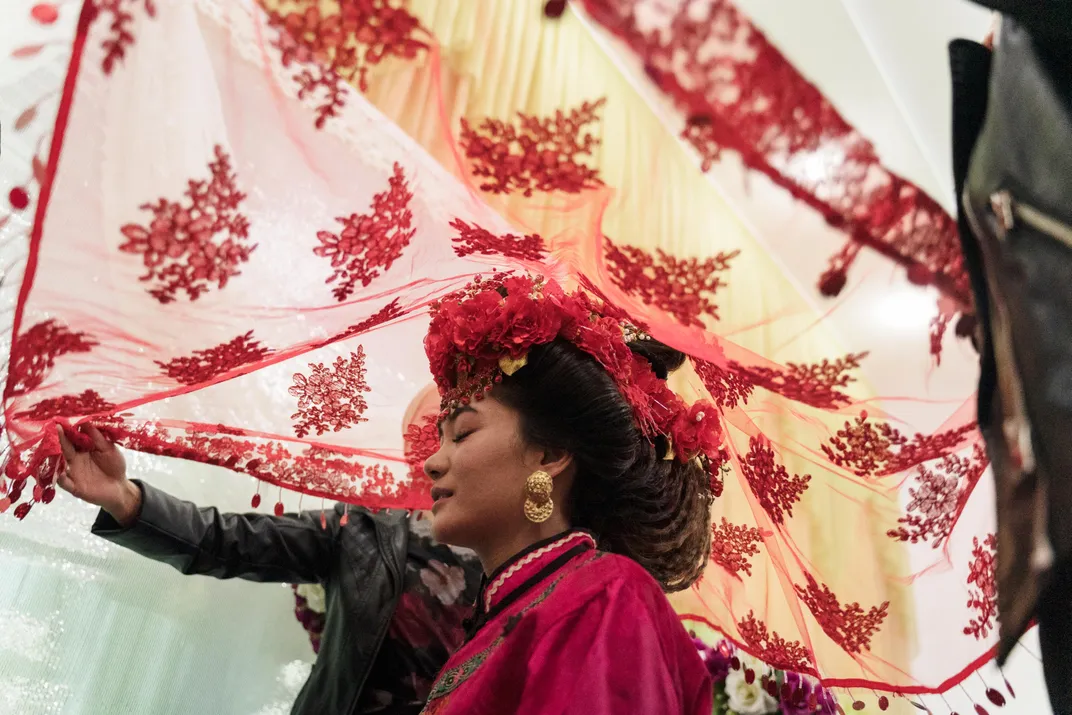 A bride wearing a red gown with a veil hovering over her head