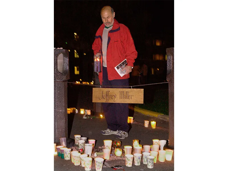 A man in a red jacket looks down at candles surrounding a sign that says 'Jeffrey Miller'