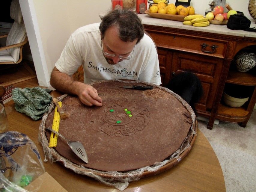 Eric Hollinger sitting at a small, wooden table bent over a large round slab of solid milk chocolate.  