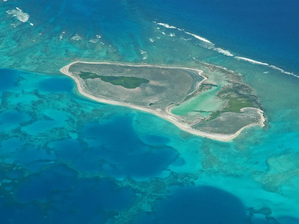An aerial image of an atoll in the Pacific Ocean. The water is marbled green and blue, and there is a blob-shaped island in the middle. It has sandy banks with green (likely plants) on the island. There are several large lagoons inside as well. 