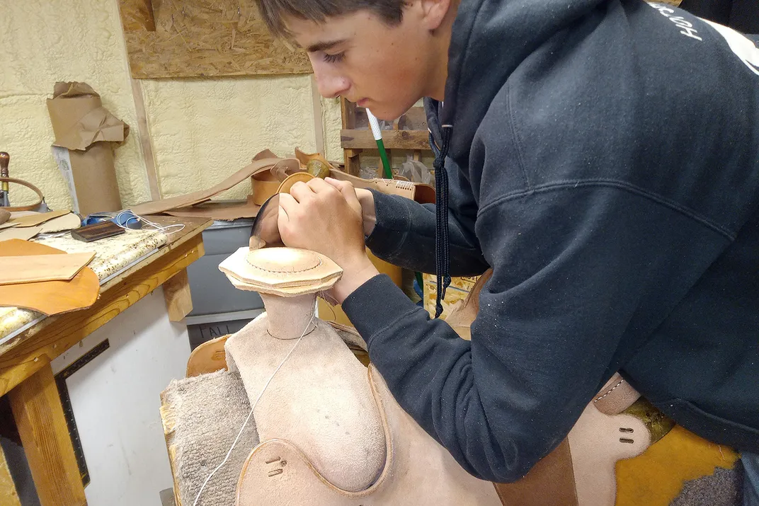 A teenage boy in a gray sweatshirt leans over a leather saddle in the making.