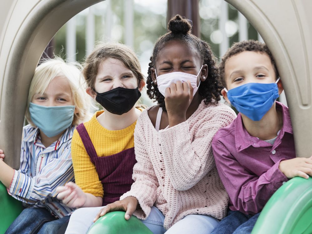 A diverse group of masked children sit at the top of a slide at a park