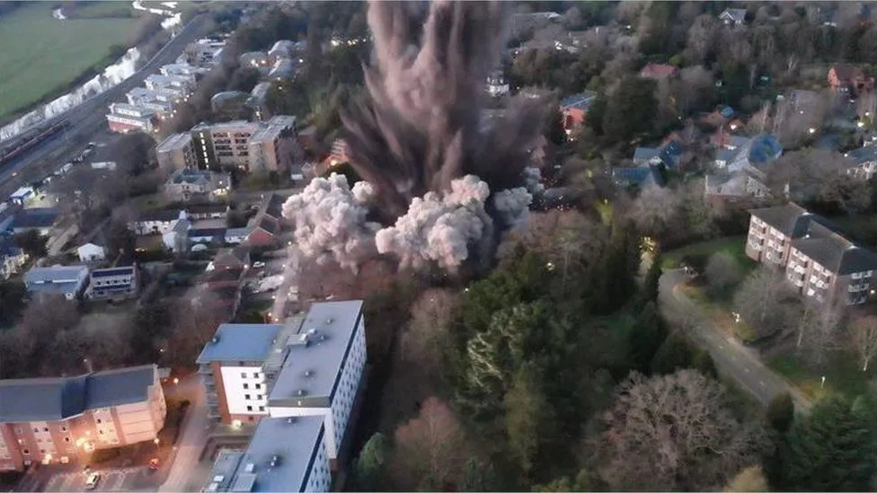 An aerial view of a huge explosion taking place in the middle of a neighborhood, with dust and smoke clouds emerging from buildings and surrounded by green trees