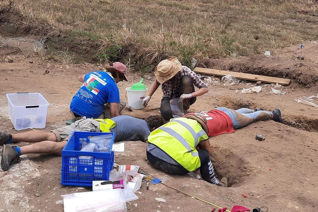 A group of four people clad in hats and reflective gear kneel and lie on their stomachs, peering into a hole dug into the dirt