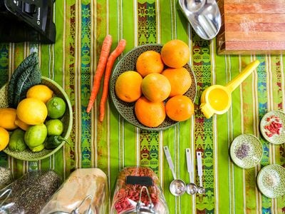 Bowls of citrus fruits including lemons, limes, and oranges, are arranged on a striped, bright green table cloth. Behind the bowls are jars filled with various superfoods including Goji berries.