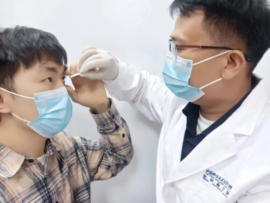 a researcher wearing a white coat, mask and gloves helps a study participant put a contact lens into his eye