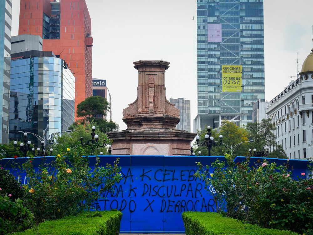 An empty pedestal stands, surrounded by tall buildings on all sides and a bright blue metal fence covered in graffiti